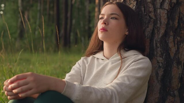 Woman thinks sitting by tree in forest. Sun rays fall on depressed female resting near trunk of plant surrounded by wild nature. Pessimistic attitude towards life