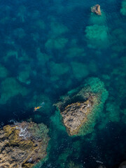 Naklejka na ściany i meble Single snorkeler in yellow swim trunks swimming in turquoise sea by the rocks in Malta aerial top down vertical