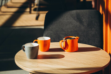 Two cups and a teapot on a table in a cafe with hard shadows. Orange teapot and cups in the...