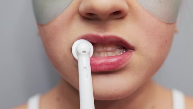 Clean and healthy habits. Dental care for kids. Unknown child little girl with under eyes patches brushing her teeth with electric toothbrush isolated on gray background