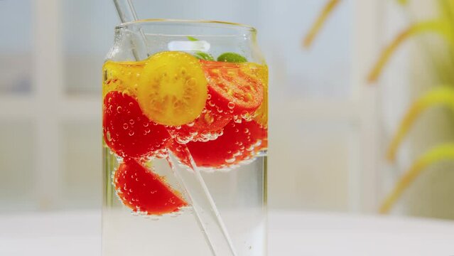 Fresh red and yellow cherry tomatoes and basil in sparkling water in glass jar with straw rotating on white table on light background close up. Detox drink, tomato juice, vegetable healthy drink bever