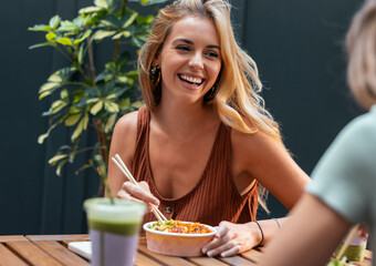 Two smiling beautiful women friends eating pokes with chopsticks while talking in the restaurant...