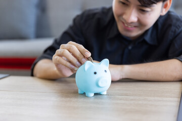 A young Asian man smiling and happy about saving by put a coins into a blue piggy bank which focus at his hand