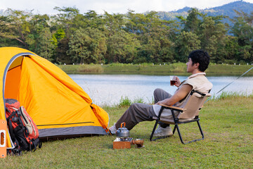 An Asian man sitting and drinking coffee near camping tent beside the lake and mountain in the sunshine morning with backpack and mobile solar cell