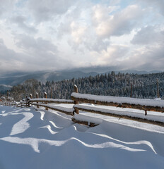 Picturesque waved shadows on snow from wood fence. Alpine mountain winter hamlet outskirts, snowy path, fir forest on far misty and cloudy hills.
