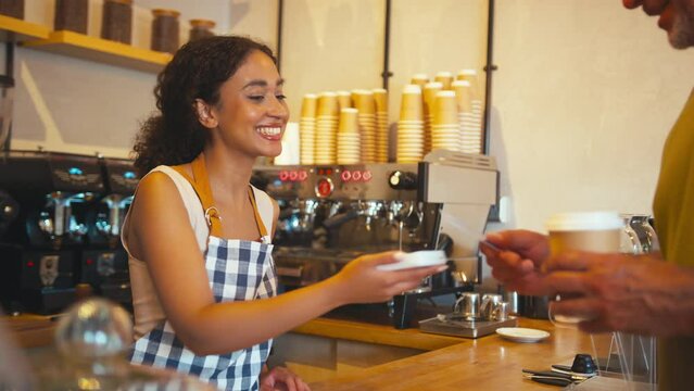 Woman wearing apron behind counter taking contactless card payment from customer in coffee shop or cafe - shot in real time