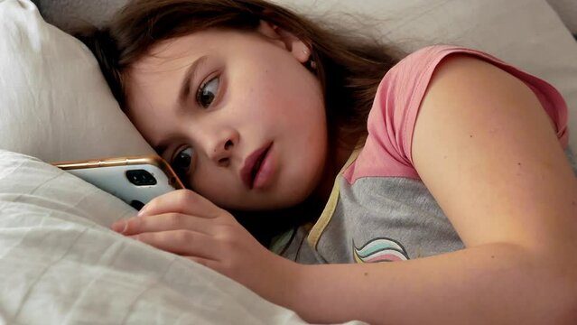 A little girl is sick, lying in bed weak and sad, looking into the frame in pajamas with disheveled hair, watching a video on a smartphone.