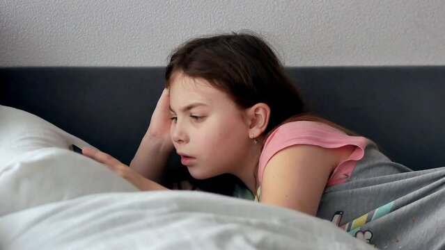 A little girl with a phone is lying in bed in the morning in pajamas with disheveled hair watching a video on her smartphone. Weekend relaxation.