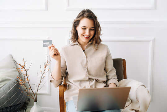 Cheerful millennial woman joyfully types on her laptop, seamlessly using a credit card for online shopping, creating an image of convenience and enjoyment in the digital retail experience at home.