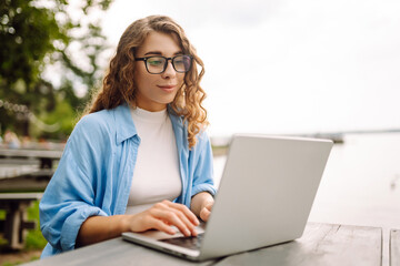 Young woman in stylish clothes sitting at a table outdoors  working on a laptop with a beautiful view of the lake. Freelancing concept. Online education.