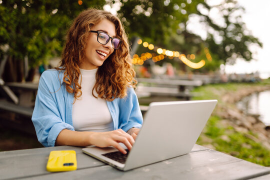 Young woman in stylish clothes sitting at a table outdoors  working on a laptop with a beautiful view of the lake. Freelancing concept. Online education.