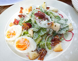 Focus on Caesar salad with crispy bacon and Boiled eggs, cut in half. Served on white plate.