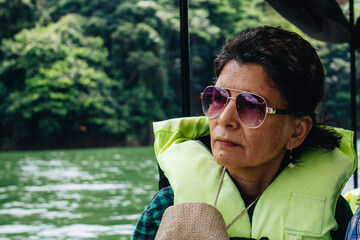 senior pensioner woman with glasses and hat with life jacket sailing on a river in a boat on vacation