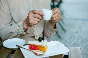Obraz na płótnie Canvas Elderly women's hands holding a cup of coffee, tea, drink outside. Senior woman, grandmother sitting at a table in a cafe terrace, with dessert, cake in the plate. Elderly concept, care, wrinkled skin