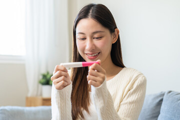 Happy thankful asian young woman hand holding pregnancy test after result with positive, surprised glad girl sitting on couch living room at home. Pregnant of ovulation fertility, motherhood concept.