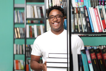 Portrait of a happy african male librarian carrying a stack of new books for readers. Dark-skinned guy in white t-shirt on the background of shelves with art books smiling cheerfully - obrazy, fototapety, plakaty