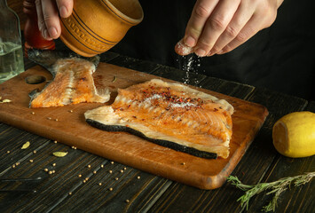 The cook adds salt to the fish steak before baking with spices and rosemary. Concept of cooking red fish on the kitchen table.