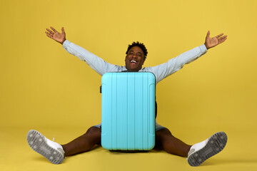 Cheerful African-American man sits behind a large suitcase with his arms and legs apart....