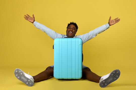Cheerful African-American man sits behind a large suitcase with his arms and legs apart. Enthusiastic happy guy traveler posing on yellow background sitting with a large travel suitcase