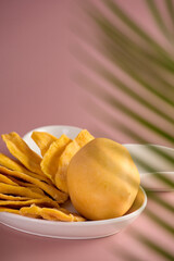 A whole mango and dried mango slices are laid out on a white plate. Mango can be seen through a tropical leaf on a pink background.