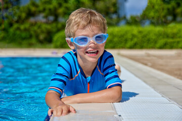 Happy Caucasian boy lies smiling on the edge of an outdoor pool. Blonde cheerful child in swimming...