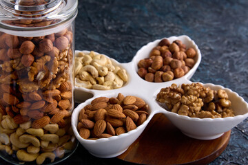 Glass transparent jar holds a mixture of different dried nuts next to a beautiful white plate on the table. Beautiful layout of nuts on a gray background in white dishes and a glass jar