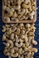 Large fresh cashew nuts, laid out on a textured gray background. Delicious cashew nuts are scattered on the table and in a wooden bowl
