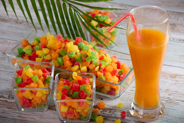 Bright sweet candied fruit is laid out in glass bowls on table, in tall glass freshly squeezed orange juice. Full glass delicious freshly squeezed juice is on table along with candied fruits in dish. 