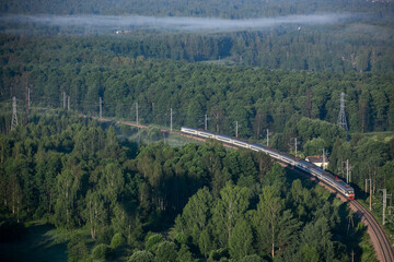 Fototapeta na wymiar An electric train moves through the forest. Power lines. Summer day. Foggy morning.