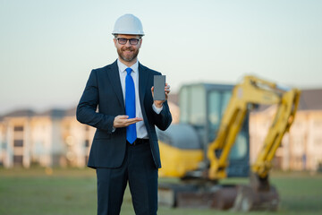 Successful construction business owner. Construction worker in suit and helmet near excavator. Confident construction owner in front of construction site. Civil engineer worker.