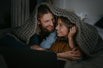Young loving couple covering with cozy warm plaid and looking at laptop while lying in bed at home
