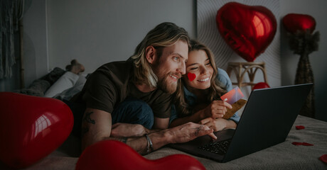 Happy loving couple looking at laptop and using credit card while shopping online at Valentines day - obrazy, fototapety, plakaty