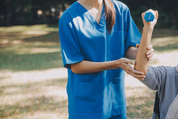 Naklejka premium Young nurse or physiotherapist in scrubs helping a happy retired old woman do fitness exercises with light weight dumbbells at home. Concept of physiotherapy for seniors