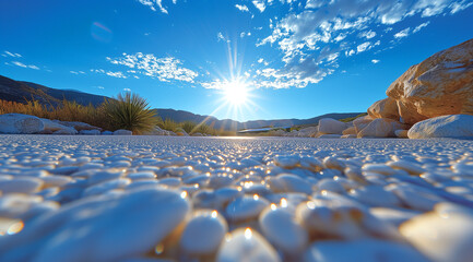 Obraz premium low angle view of a bright sun in a blue sky with clouds, over a desert landscape with white pebbles, plants, and large rocks