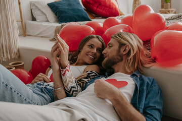 Young loving couple holding Valentines cards while leaning on bed surrounded with red balloons