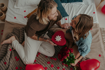 Top view of romantic young man giving a red gift box to his girlfriend while sitting on bed together