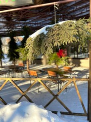 a street umbrella made of green fir branches with a red bow on the background of tables and chairs in the park in winter in a cafe against the background of snow