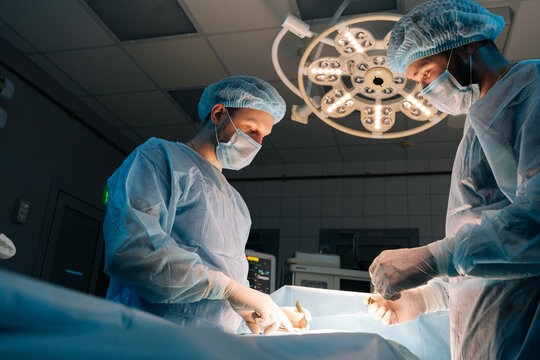 Low-angle View Of Two Skilled Male Surgeons Wearing Sterile Gloves, Surgical Uniforms And Masks Processing Surgical Operation In Dark Operating Room Modern Hospital Emergency Department.