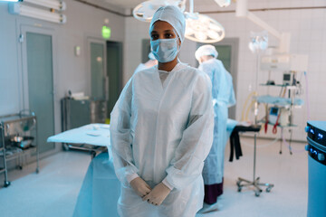 Portrait of African-American female nurse standing posing looking at camera in operating room with light modern interior. Diverse team of surgeons performing operation on background