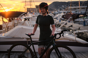 Female cyclist wearing cycling kit, glasses and helmet standing with her bike during sunset on...
