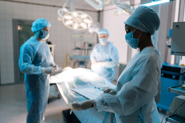 Group of multiethnic surgeons and nurse wearing on disposable gloves as hygiene before surgery in hospital with modern light interior. Concept of preparing for surgical operation. - obrazy, fototapety, plakaty