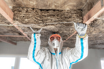 The ceiling insulation with a glass wool. The Worker dressed in a protective coveralls is fixing a...