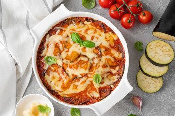 Eggplant casserole with cheese and tomato sauce in a white baking dish on a gray concrete background with ingredients for cooking. Vegetarian healthy food.