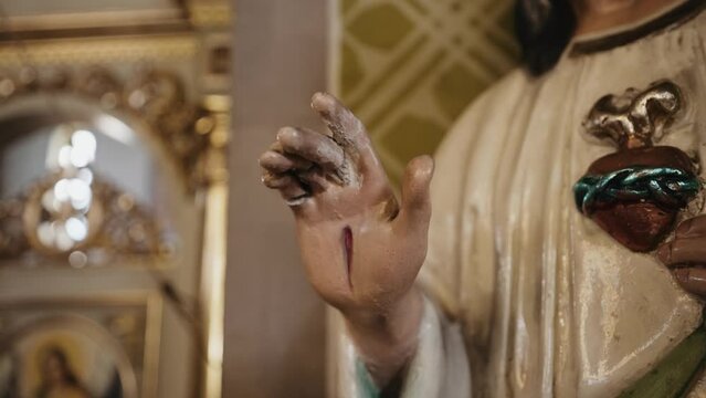 Close-up of a religious statue's hand with stigmata and a sacred heart adorned with a crown of thorns. The background is softly out of focus, highlighting the statue's details.