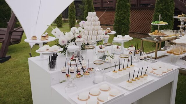 Elegant dessert table at an outdoor event with variety of sweets. Includes macaron tower, assorted pastries, and cupcakes. White and floral decor create luxurious setting, weddings or upscale parties.