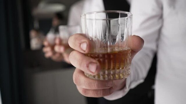 hand holding a glass of whiskey. The person is wearing a white shirt with a cufflink visible. Another hand with a glass is in the background. The focus is on the glass in the foreground, suggesting 