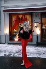 beautiful woman with blond hair in elegant fur coat and accessories posing near shop window decorated for Christmas