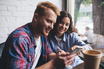 Portrait of cheerful Caucasian blogger smiling at camera while bearded boyfriend searching website on modern smartphone technology, happy couple in love using cellphone device for social networking - obrazy, fototapety, plakaty