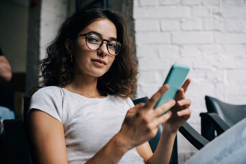 Portrait of attractive female with smartphone technology looking at camera during free time, millennial blogger in optical spectacles for provide vision correction holding modern mobile device