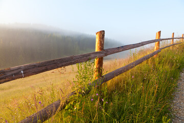 Wooden fence near the meadow on a foggy morning, sun shines through the fog. Ukraine, Carpathians.